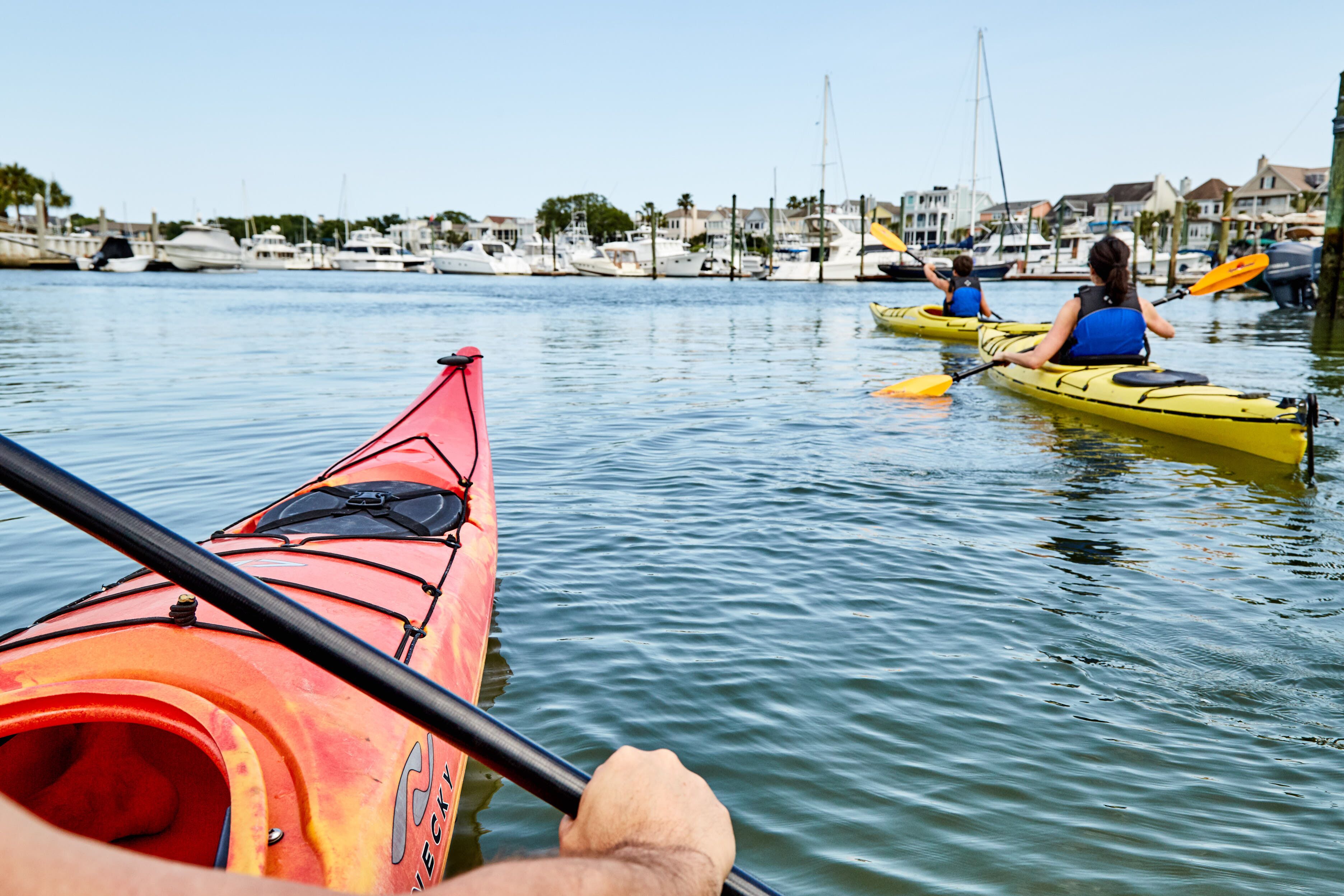 Un puerto deportivo con kayaks y barcos, agua tranquila y casas al fondo.