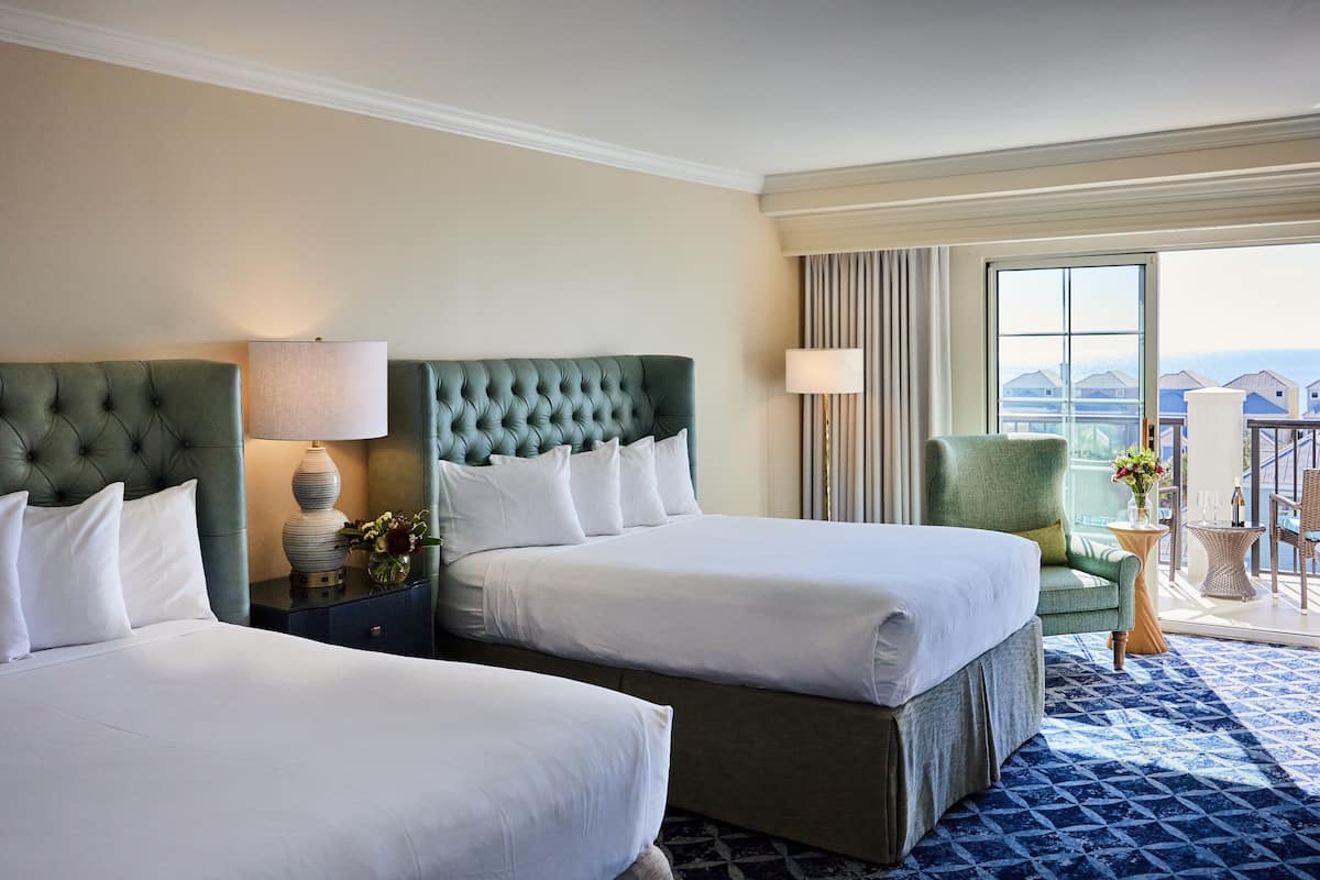 A hotel room with two beds, a green armchair, a small table with a vase, and a balcony with a view.
