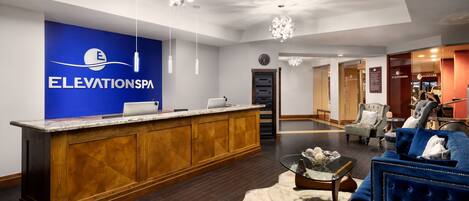 Un área de recepción con un mostrador de madera, una pared azul con el logotipo 'ElevationSpa' y una zona de estar con un sofá y sillones azules.