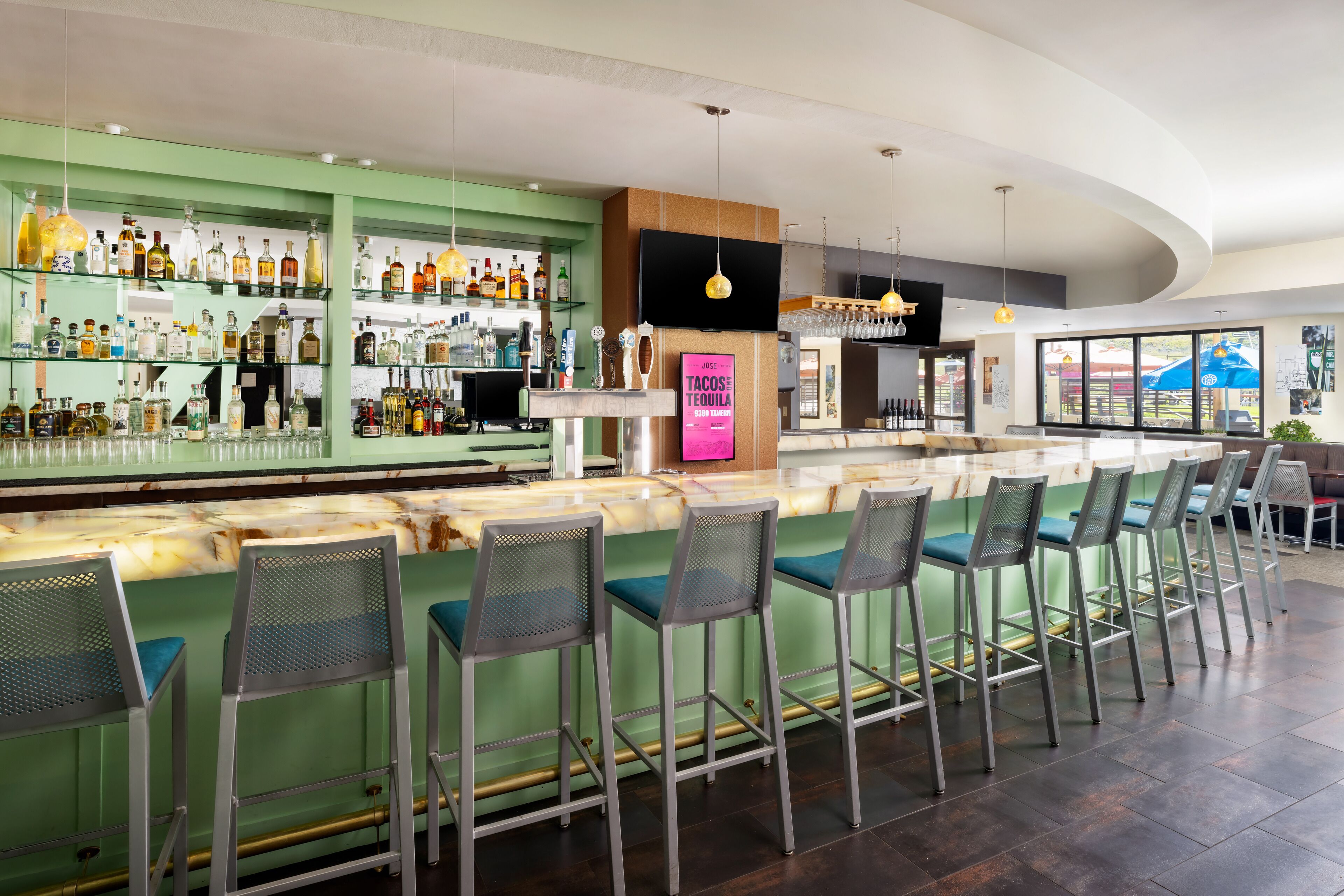 Un espace bar avec un comptoir vert, de nombreux tabourets et une variété de bouteilles et de verres exposés sur des étagères.