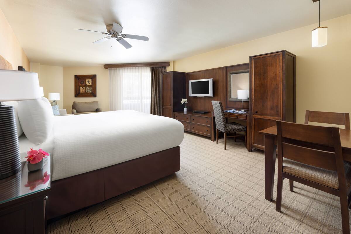 A hotel room with a large bed, a desk with a computer, a TV, and a ceiling fan.