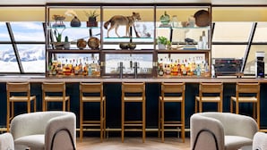 Uma área de bar com vista para montanhas, banquetas e prateleiras com garrafas e objetos decorativos.