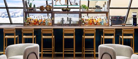 Un espace bar offrant une vue sur les montagnes, avec des tabourets de bar et des étagères remplies de bouteilles et d’objets décoratifs.