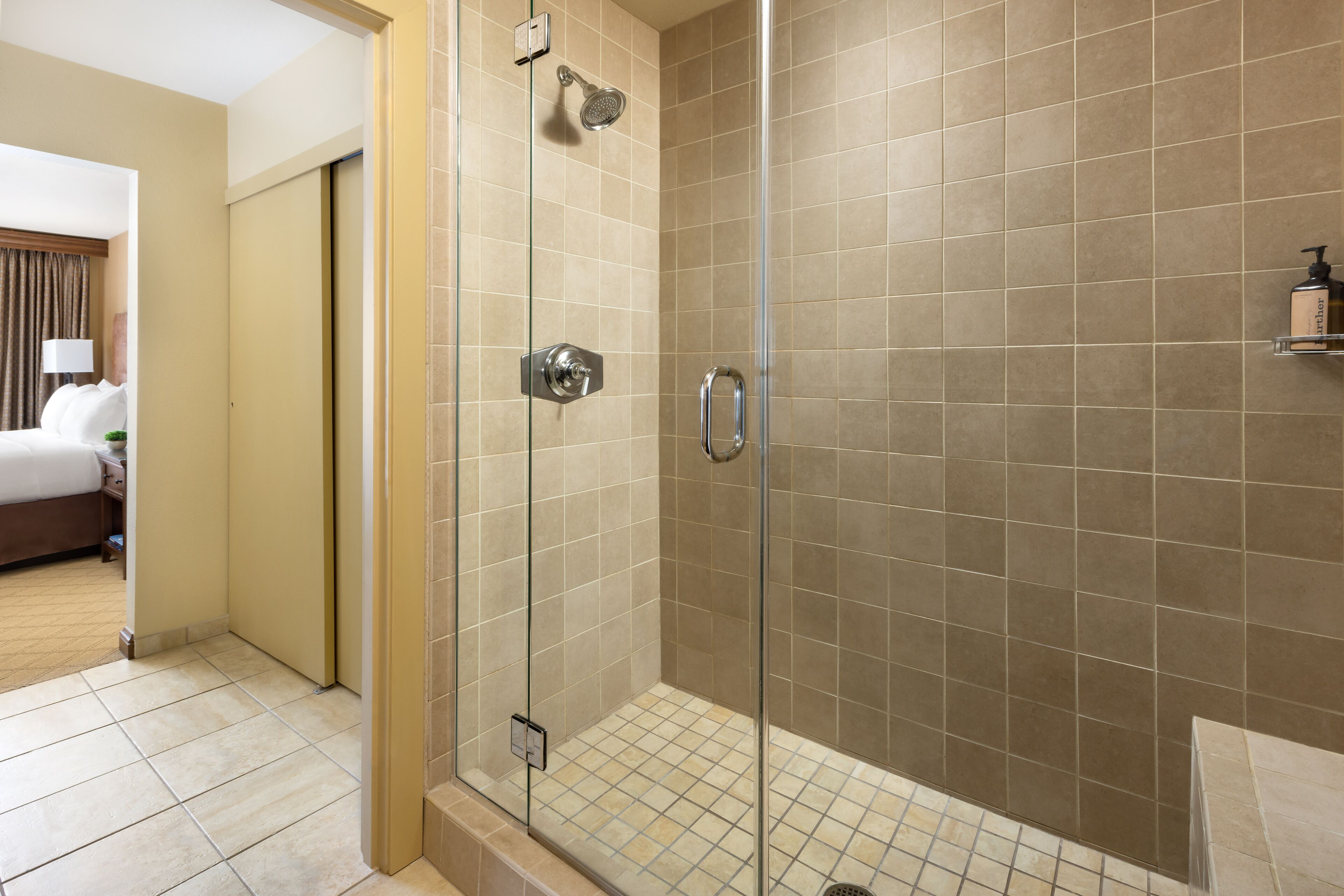 Une douche fermée par des parois de verre, avec des carreaux de couleur beige, un panneau de commande visible et une petite étagère contenant des articles de toilette.