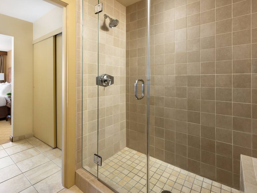 Une douche fermée par des parois de verre, avec des carreaux de couleur beige, un panneau de commande visible et une petite étagère contenant des articles de toilette.