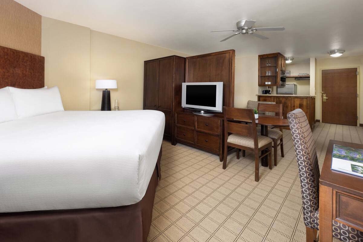 A hotel room with a large bed, a desk with a television, a dining area with chairs, a ceiling fan, and a kitchenette.