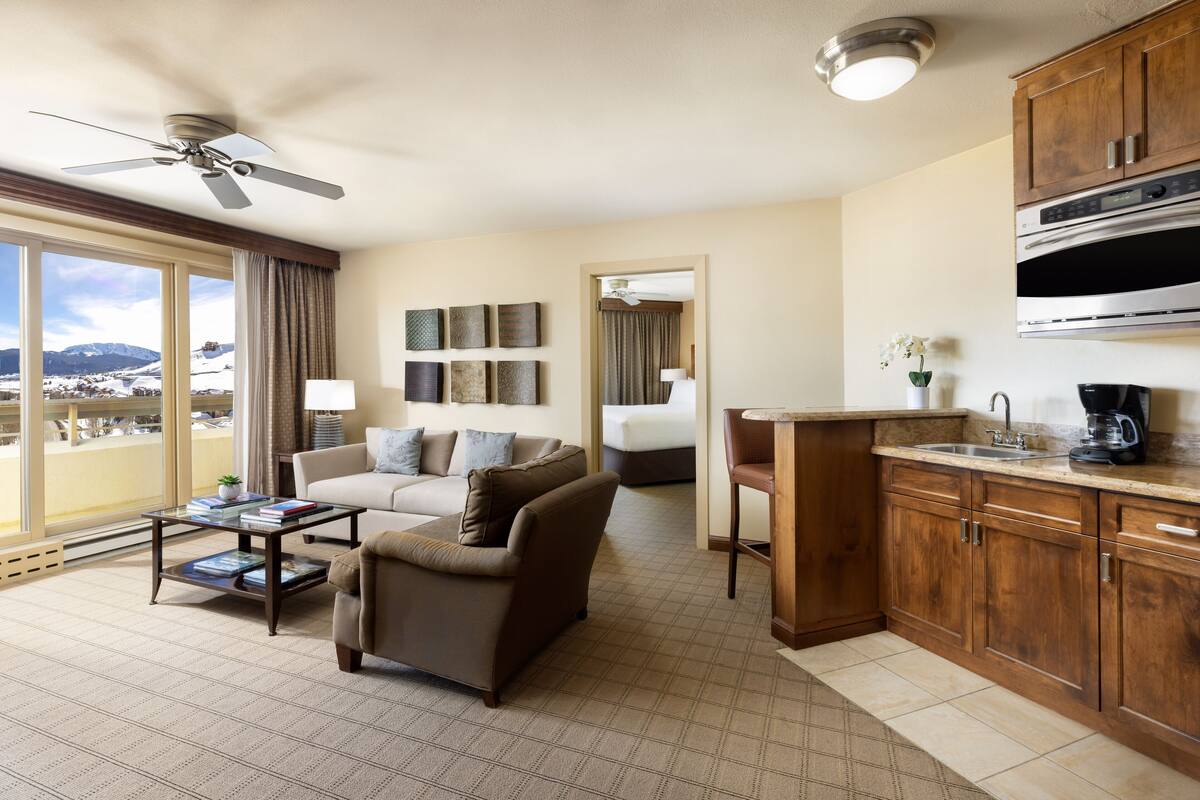 A hotel room with a living area, kitchenette, and a balcony with a view of snow-covered mountains.