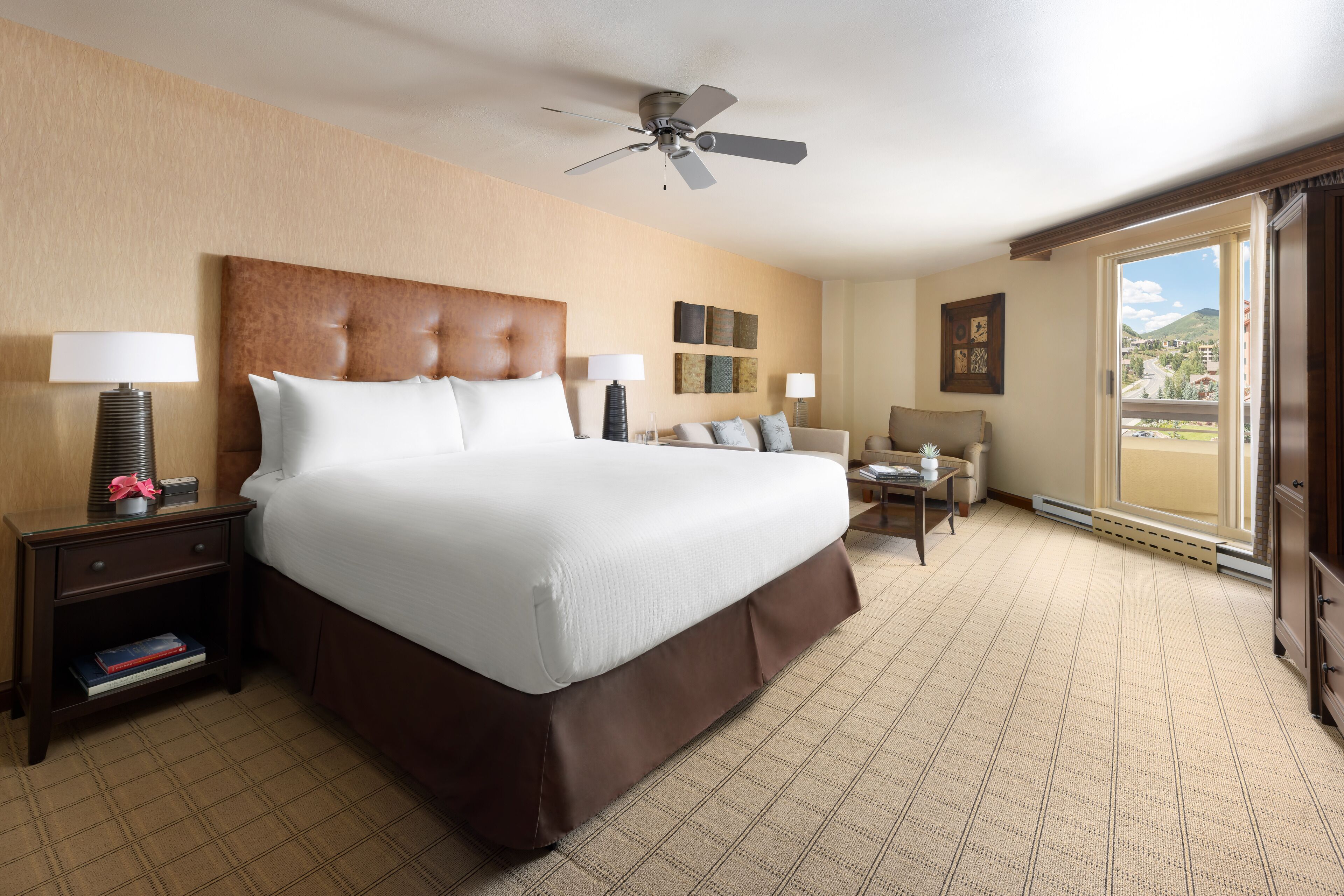 A hotel room with a large bed, a ceiling fan, a desk with a chair, and a view of the outside.