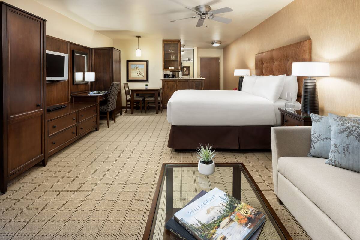 A hotel room with a large bed, a desk with a chair, a television, a minibar, and a ceiling fan.