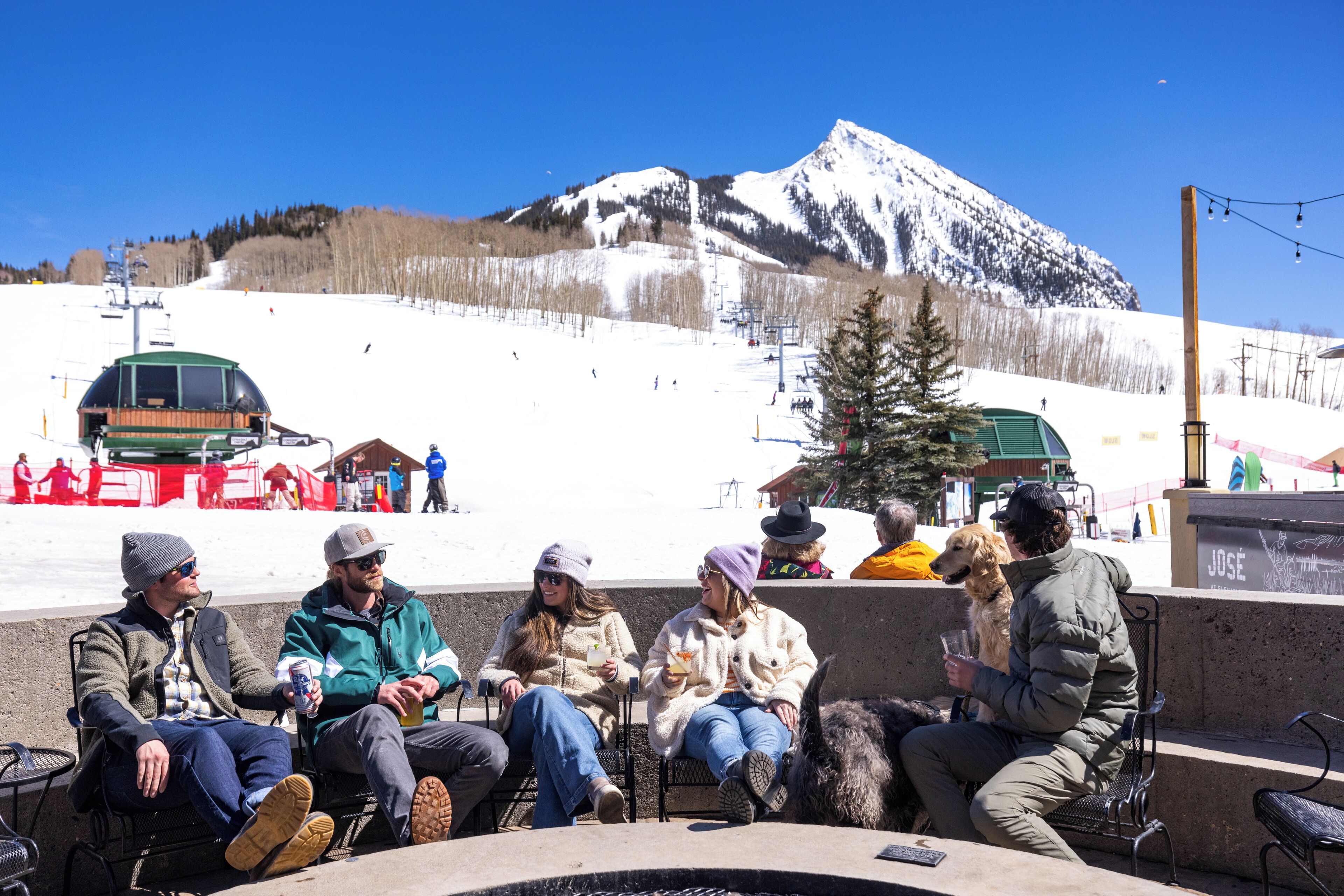 A group of people sitting around a fire pit in a snowy outdoor setting with a mountain in the background.
