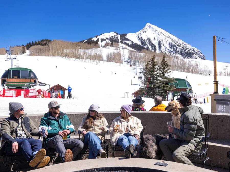 A group of people sitting around a fire pit in a snowy outdoor setting with a mountain in the background.