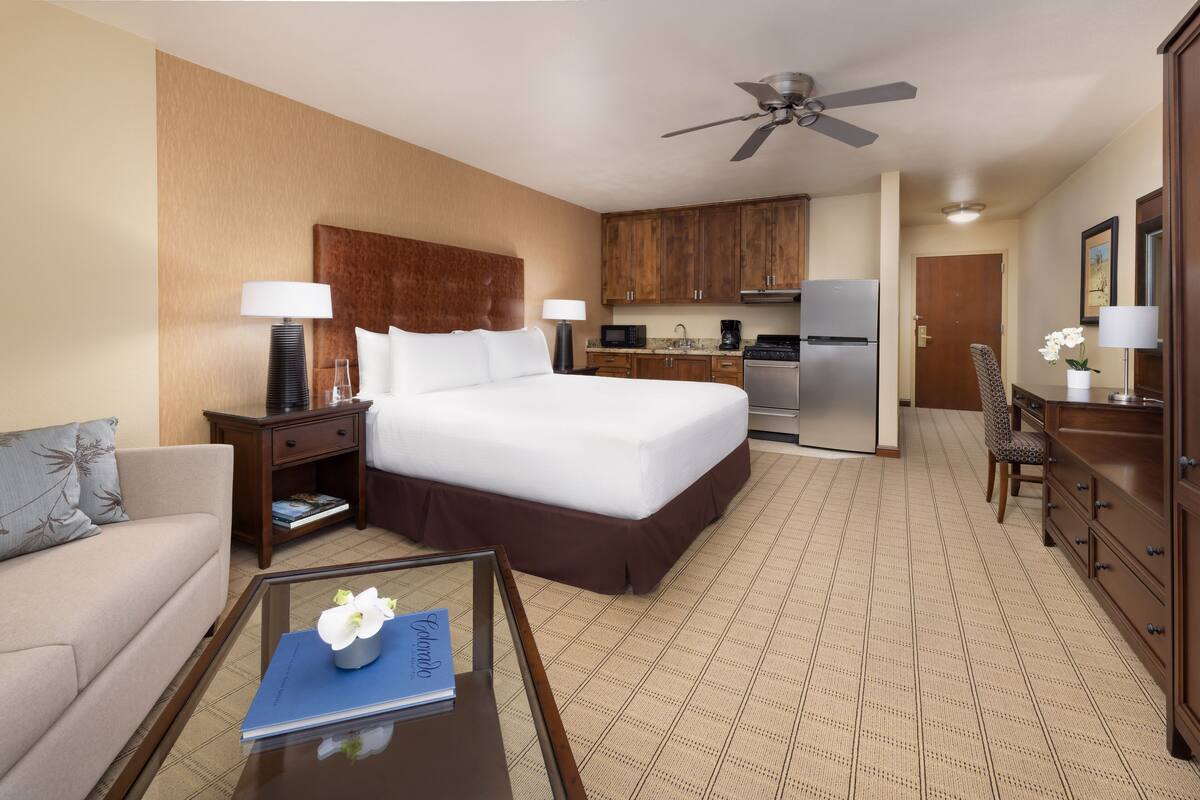 A hotel room with a large bed, a small kitchenette, a dining area, and a living space with a sofa and coffee table.