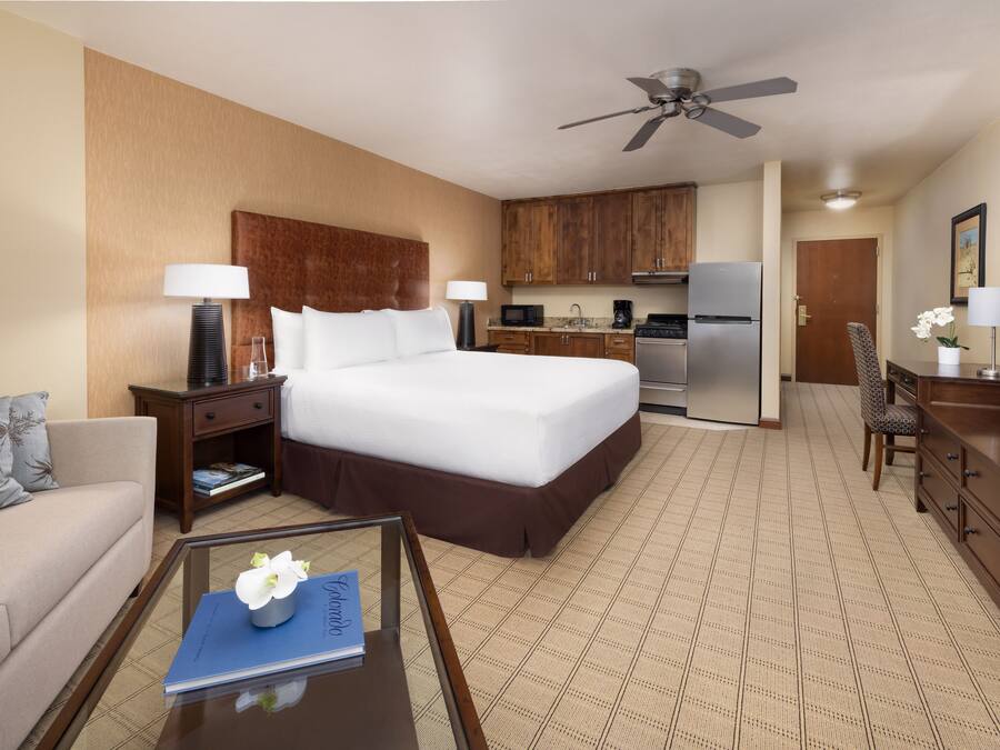 Una habitación de hotel con una cama grande, una kitchenette pequeña, un comedor y una sala de estar con sofá y mesa de centro.