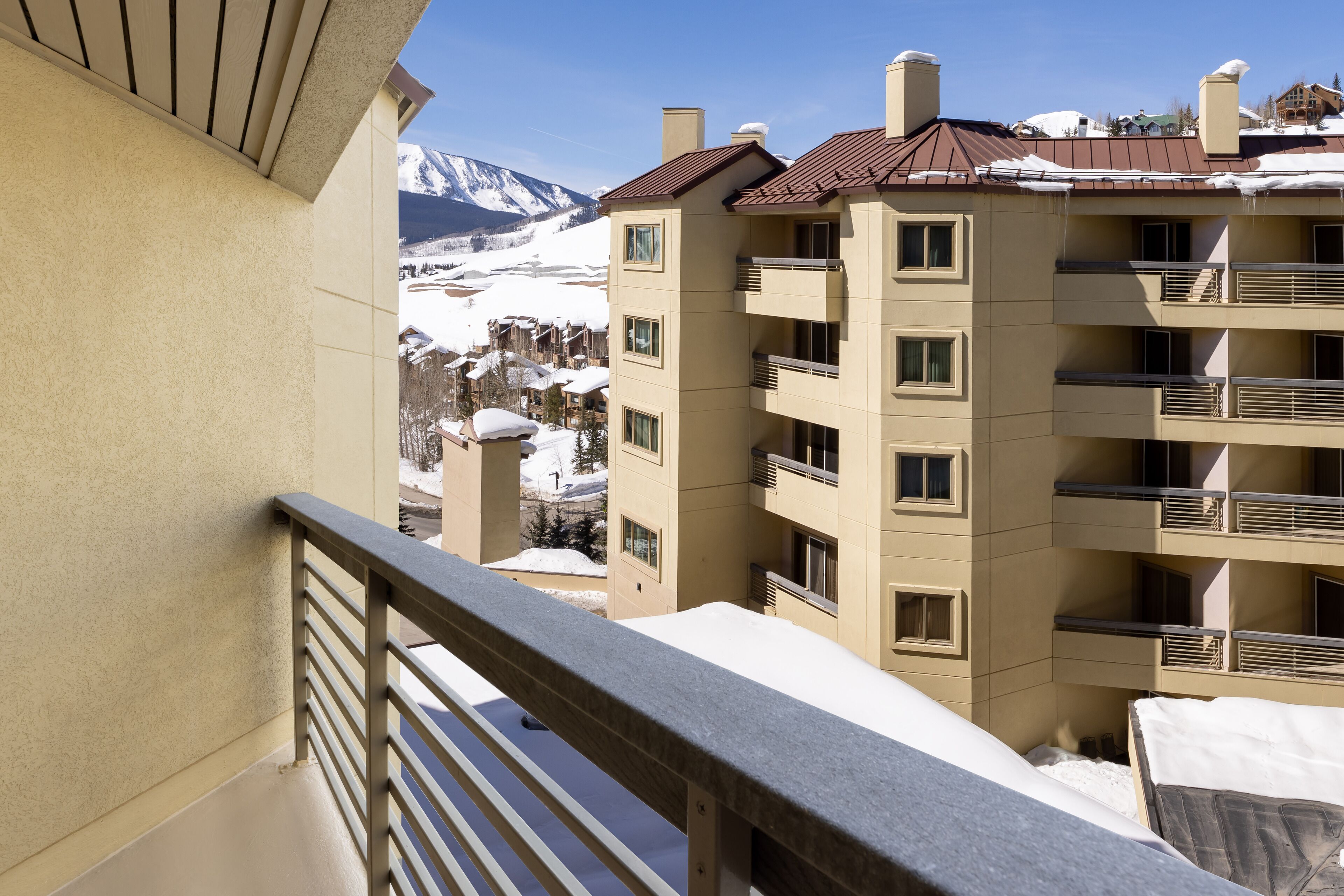 Un edificio de varios pisos con balcones, techo cubierto de nieve y montañas al fondo.
