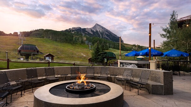 A circular outdoor seating area with a fire pit, surrounded by chairs and tables, overlooking a mountainous landscape. - Elevation Hotel & Spa (Crested Butte)