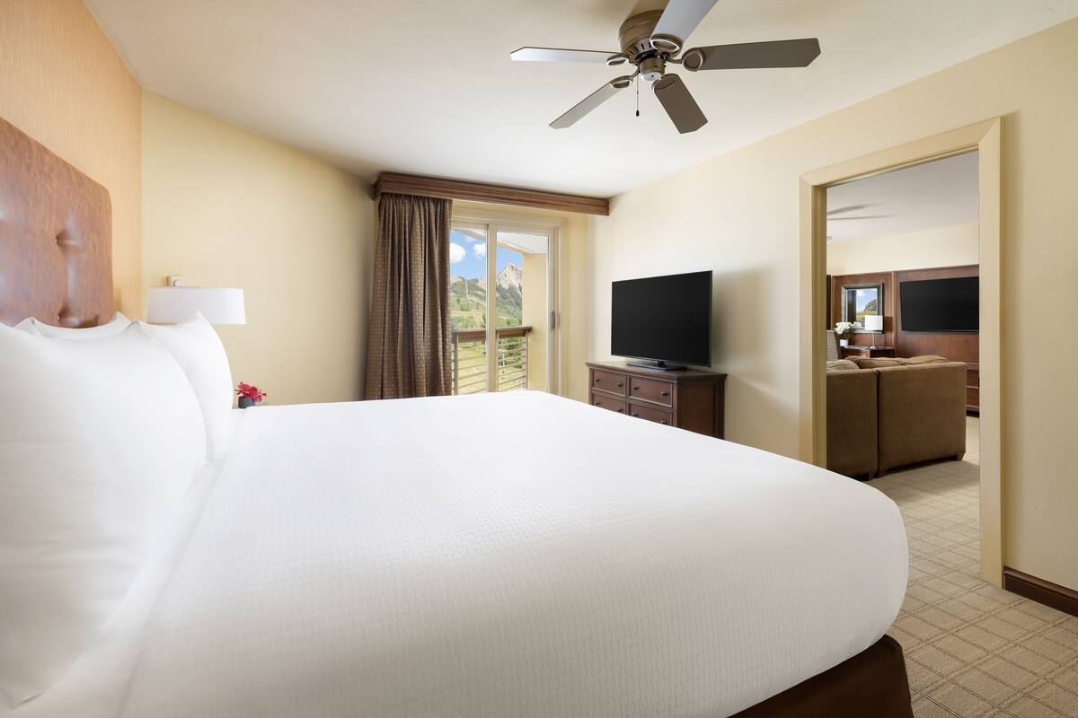 A hotel room with a large bed, a ceiling fan, a television, and a view of the outdoors.