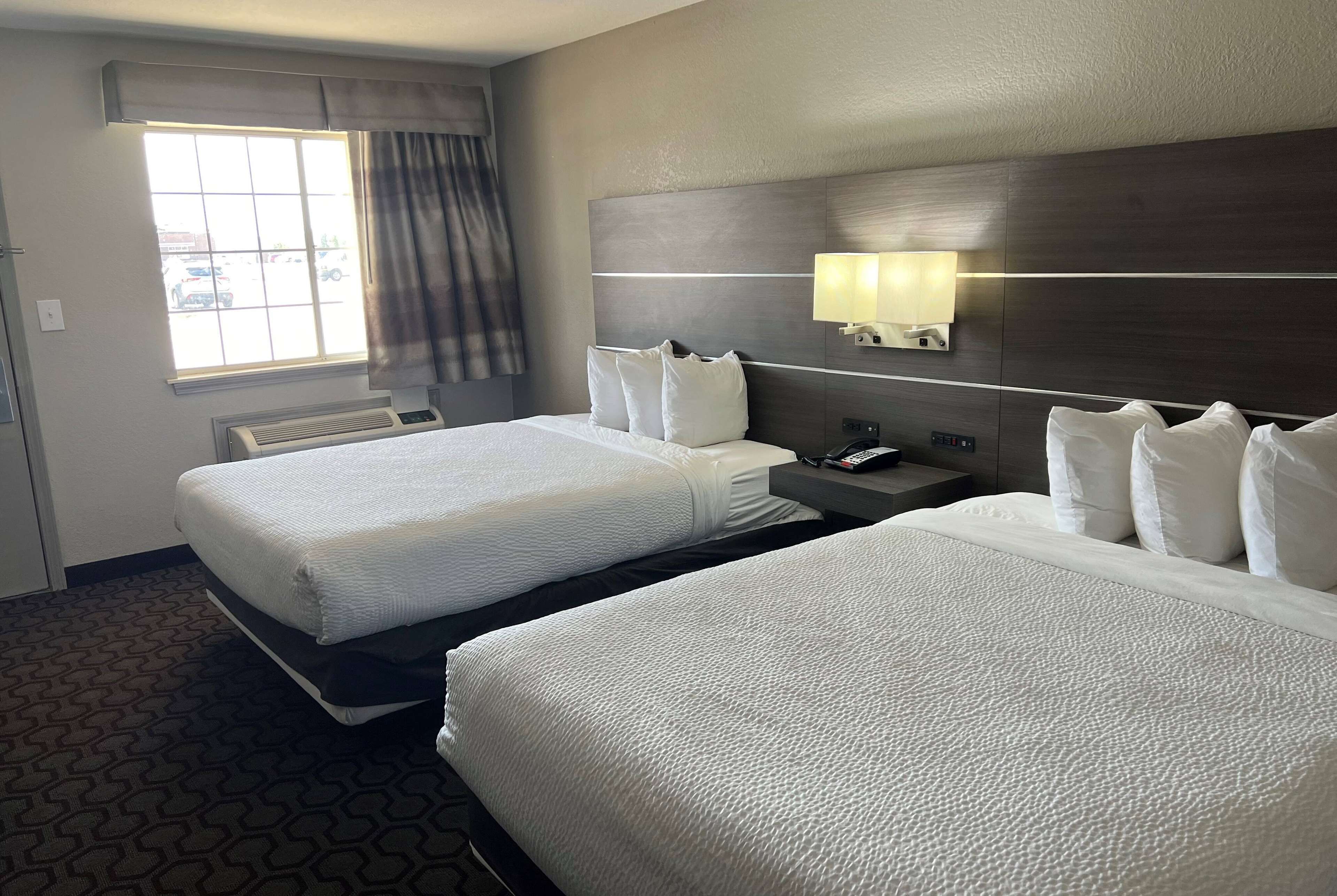 A hotel room with two beds, a window with curtains, a wall-mounted lamp, and a control panel.