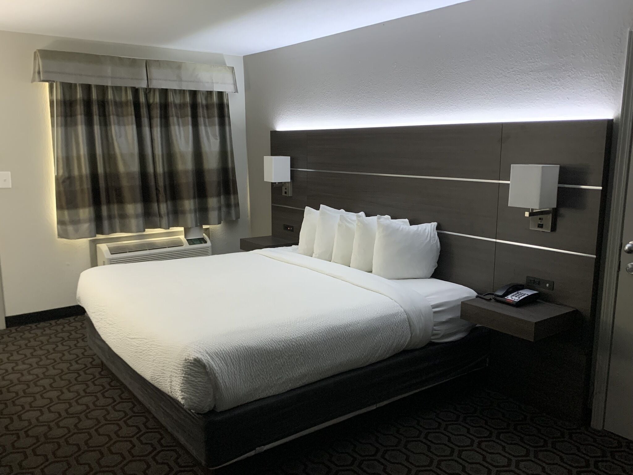 A hotel room with a large bed, a bedside telephone, a nightstand, a wall-mounted lamp, and a window with curtains.