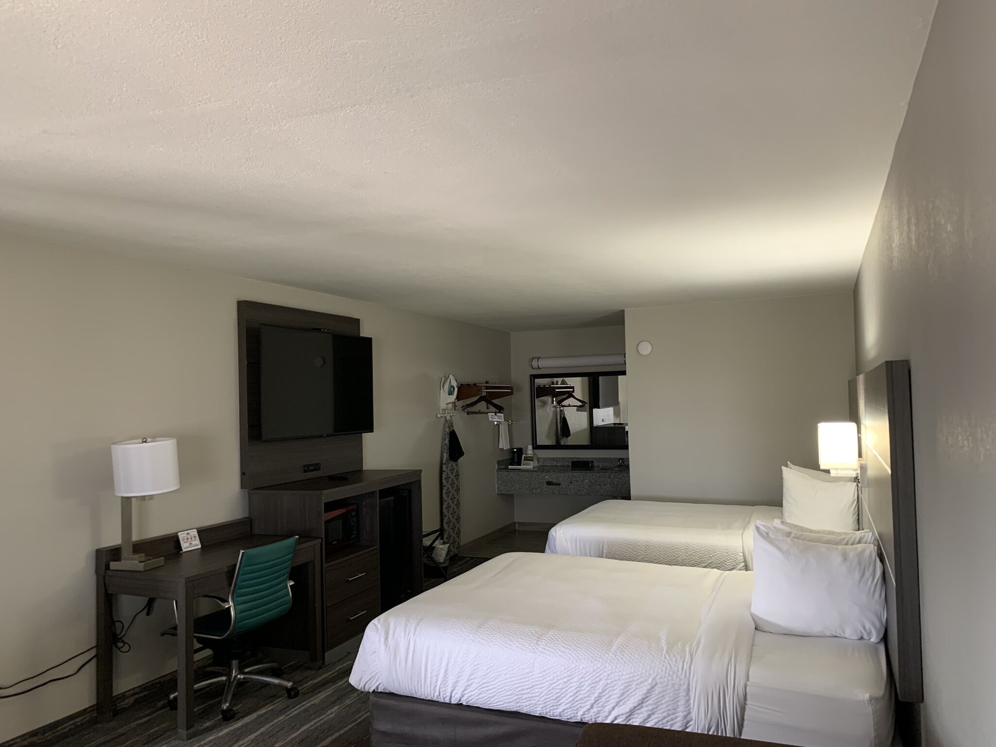 A hotel room with two beds, a desk with a chair, a television, a mirror, and a small kitchenette.