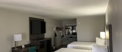 A hotel room with two beds, a desk with a chair, a television, a mirror, and a small kitchenette.