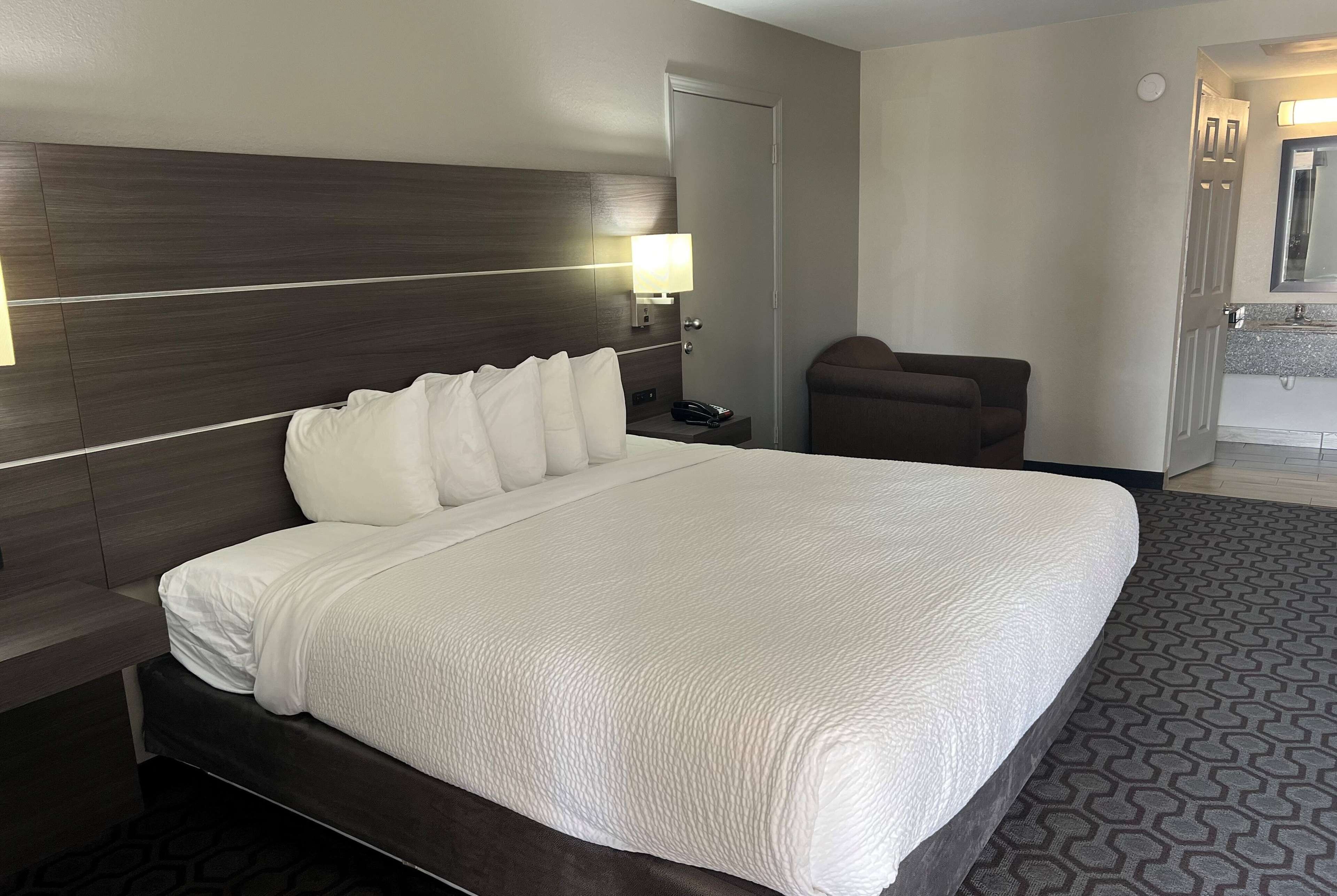 Une chambre d’hôtel avec un grand lit, une table de chevet, une lampe, une chaise et une salle de bain visible au fond.