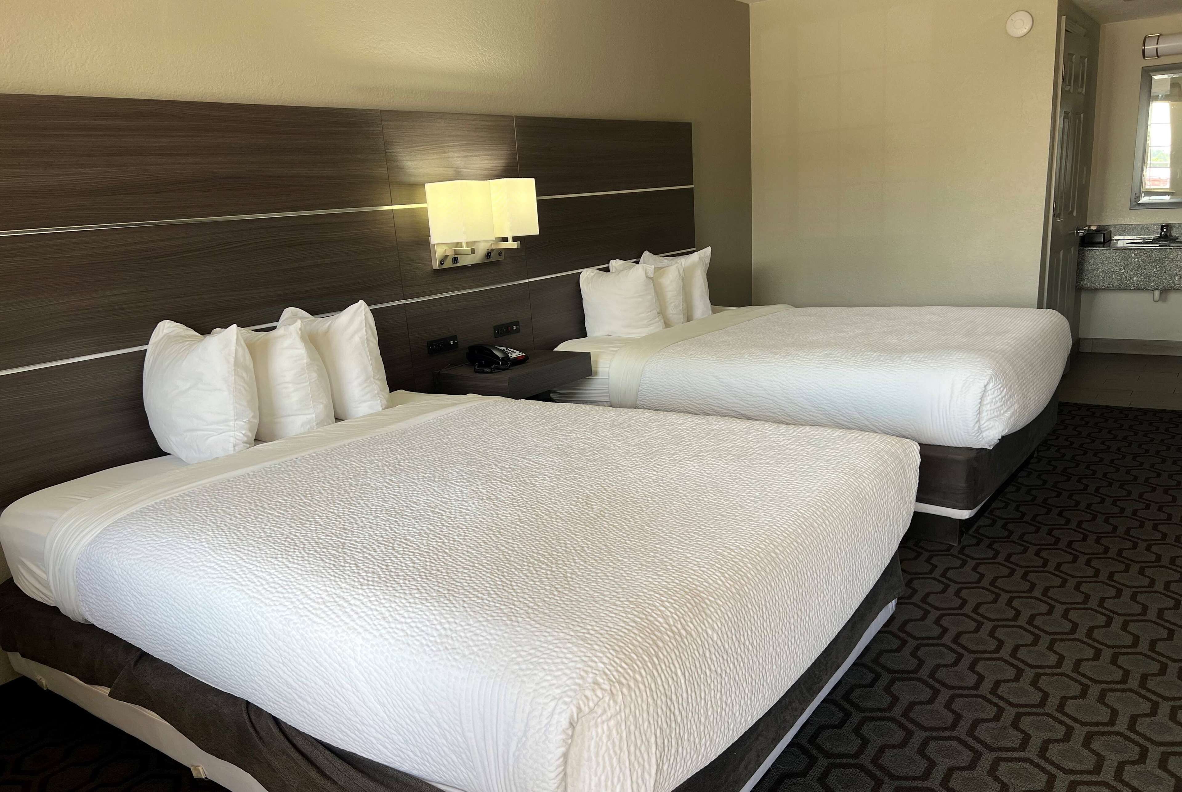 A hotel room with two beds, a nightstand, a lamp, and a bathroom with a sink and mirror.