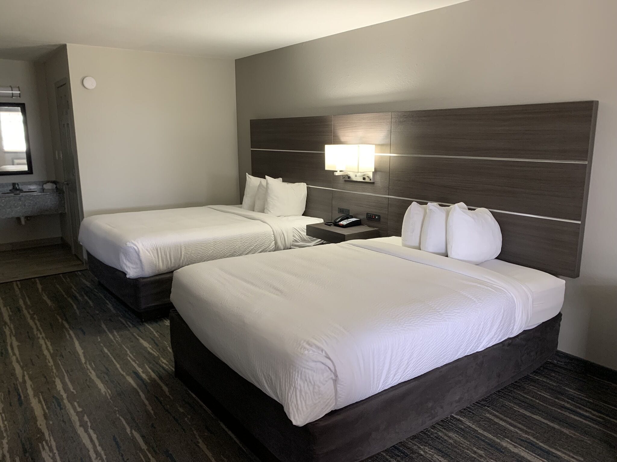 Habitación de hotel con dos camas, una mesita de noche, una lámpara y un baño visible a través de una puerta abierta.