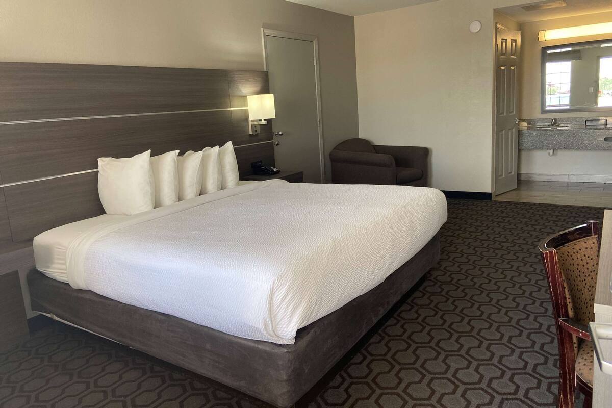 A hotel room with a large bed, a nightstand, a chair, a desk, a mirror, and a bathroom with a sink and mirror.