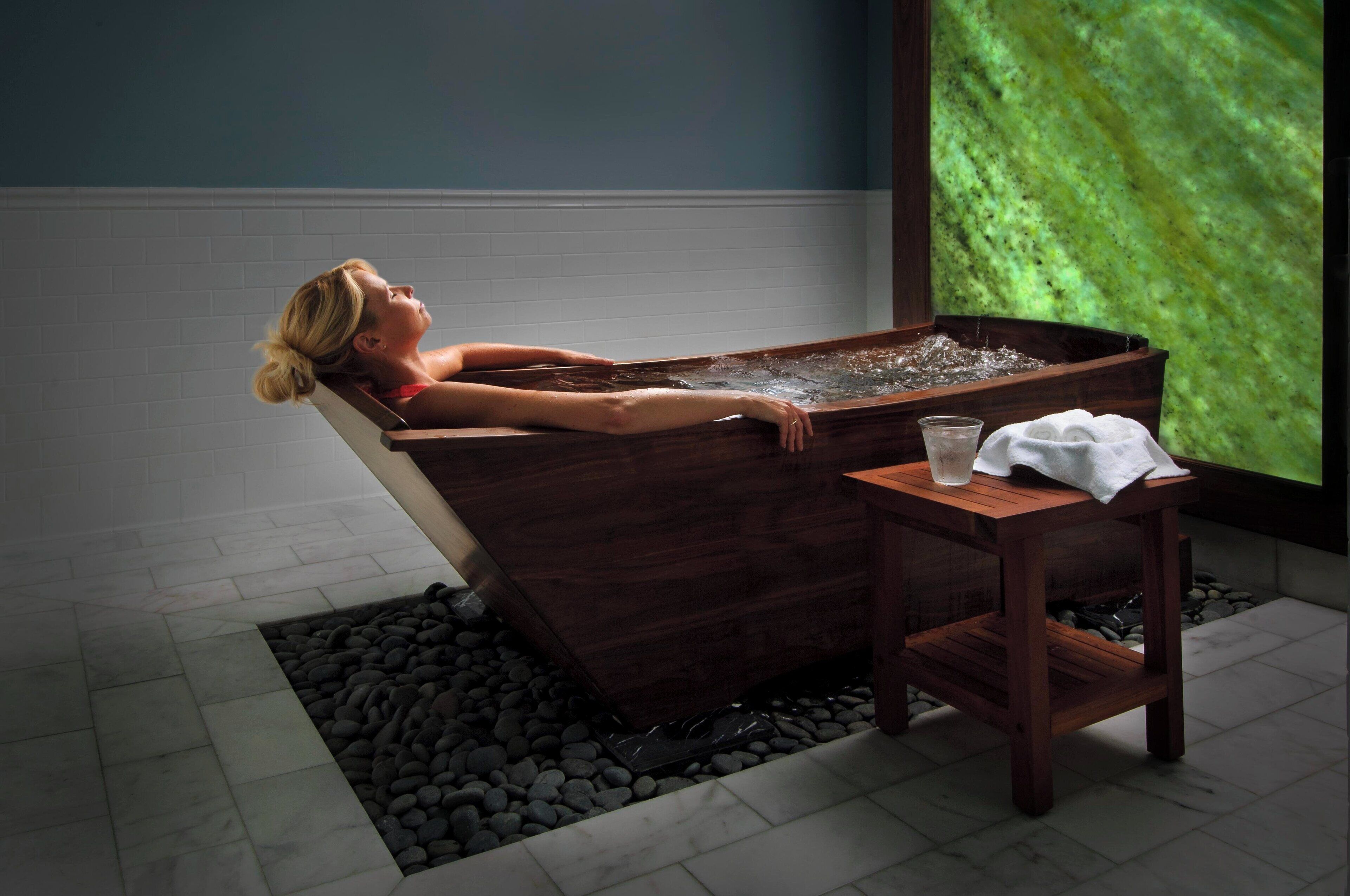 A person relaxing in a wooden bathtub with a small table beside it.
