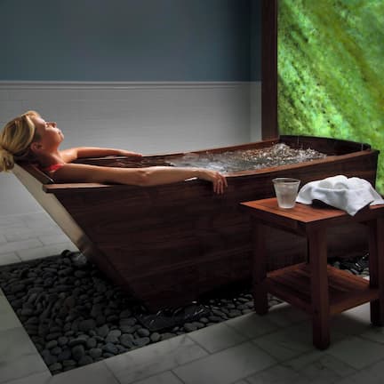 A person relaxing in a wooden bathtub with a small table beside it.