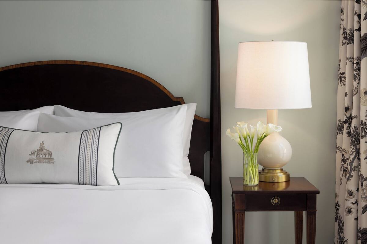 A neatly made bed with white linens and a decorative pillow, a bedside table with a lamp and flowers, and a floral curtain.