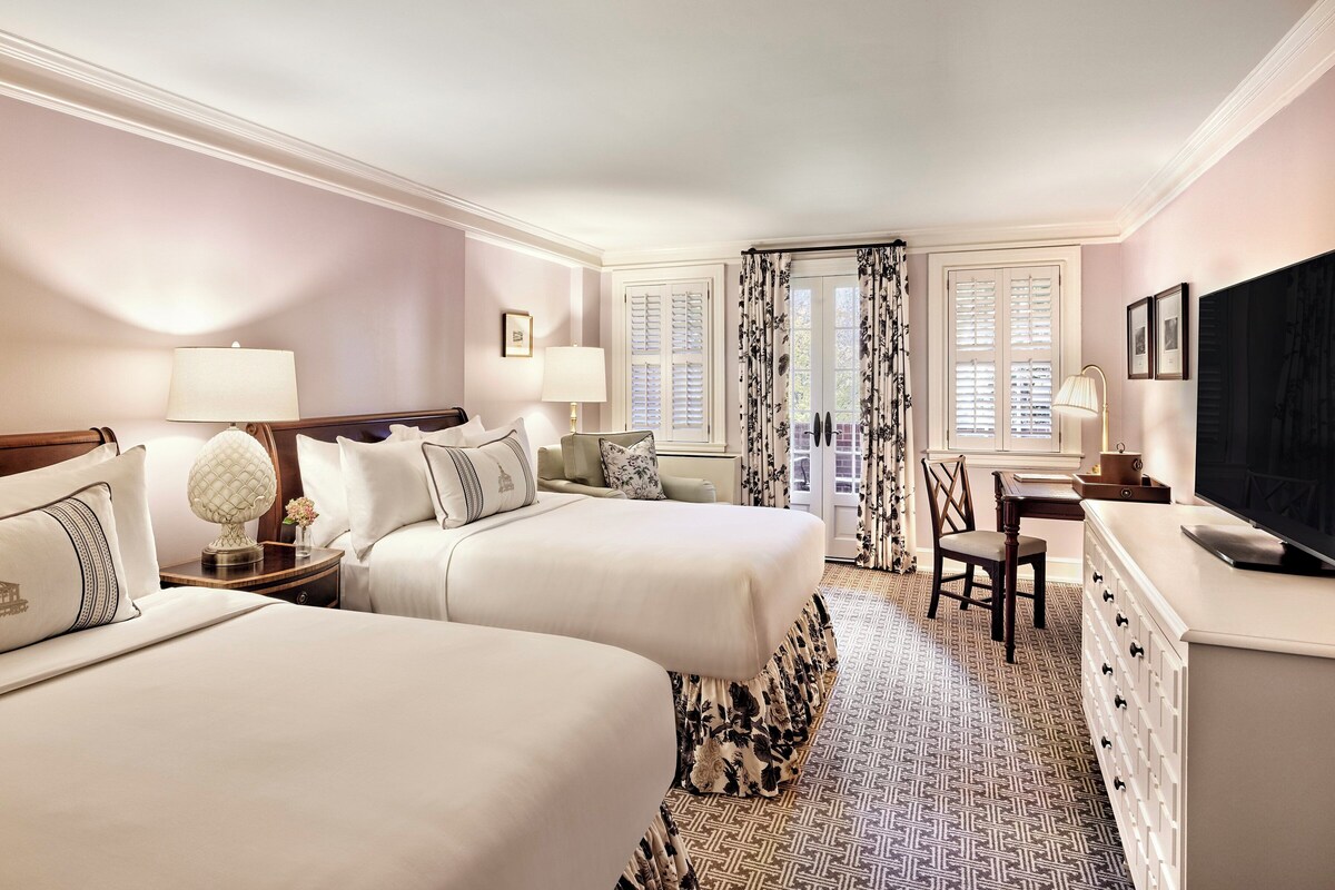 A hotel room with two beds, a desk, a chair, a television, and a window with shutters.