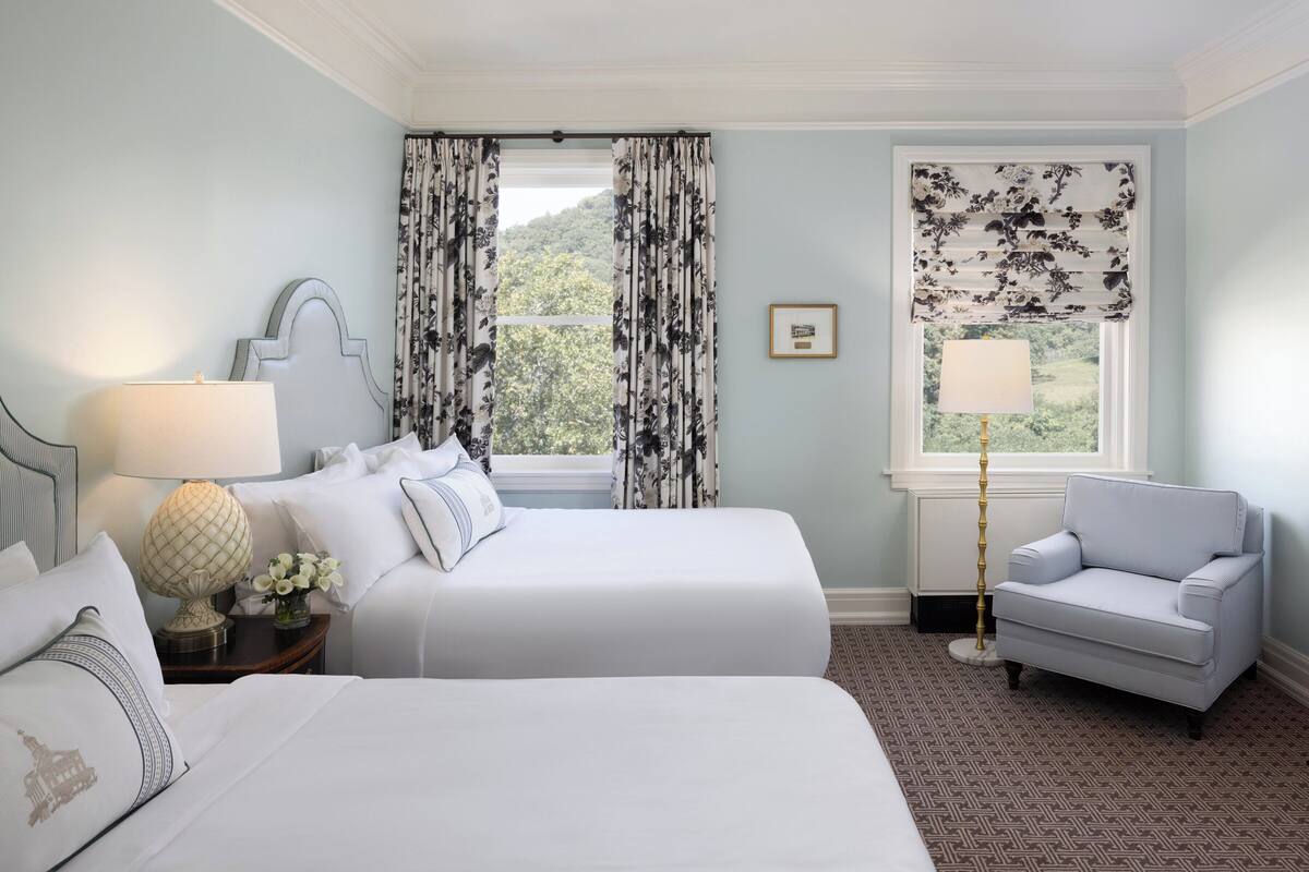A hotel room with two beds, a chair, a lamp, and a view of greenery through the window.