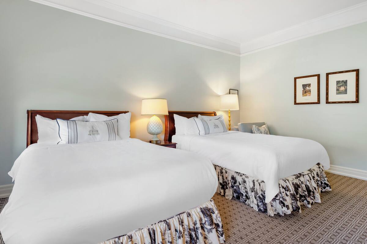 A hotel room with two large beds, each with white bedding and floral-patterned skirts. There are two bedside lamps and two framed pictures on the wall.