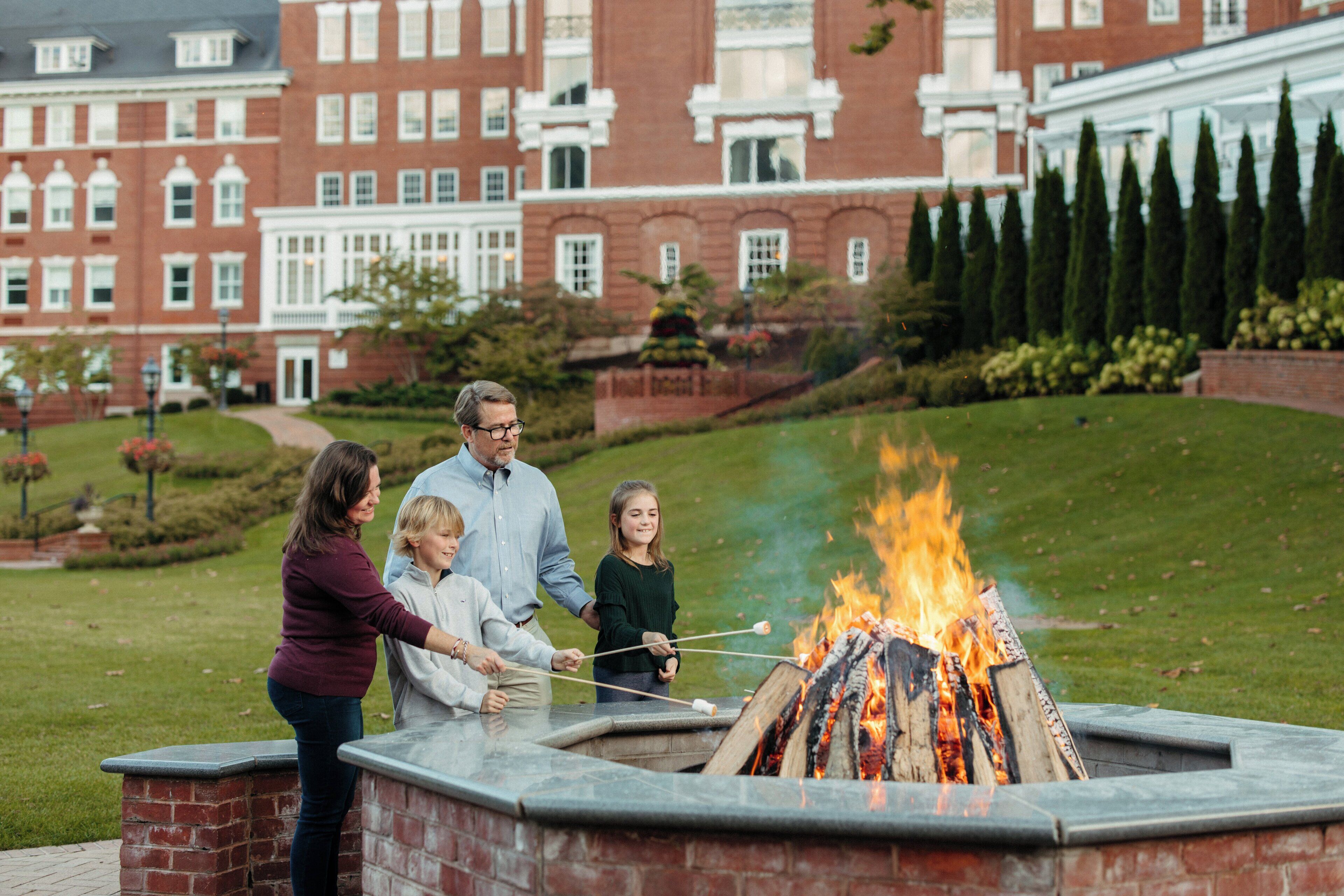A family gathered around a fire pit with a brick structure, set against a backdrop of a red brick building with multiple windows and a well-maintained lawn.