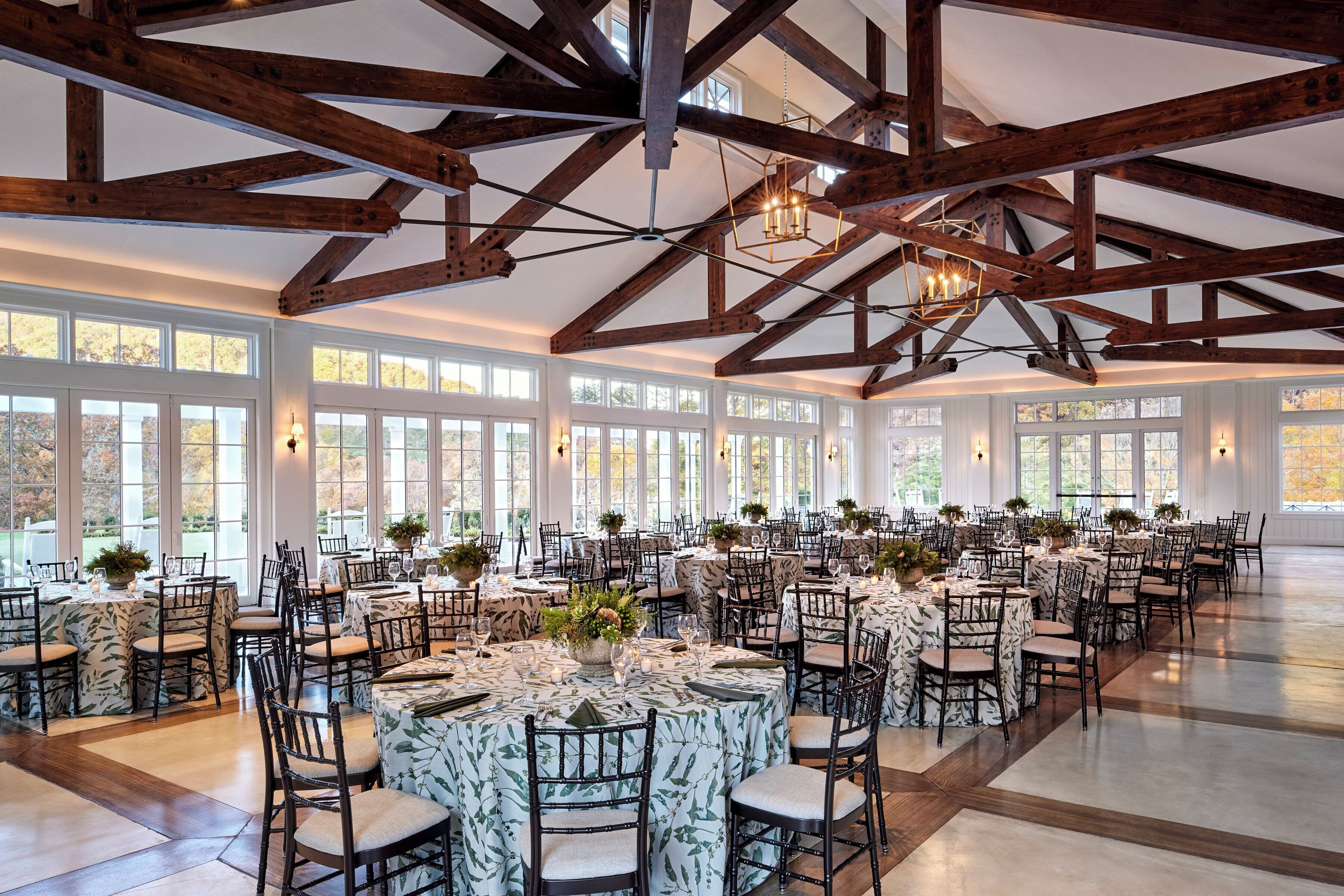 A large hall with high ceilings and exposed wooden beams, furnished with round tables and chairs, and large windows allowing natural light.