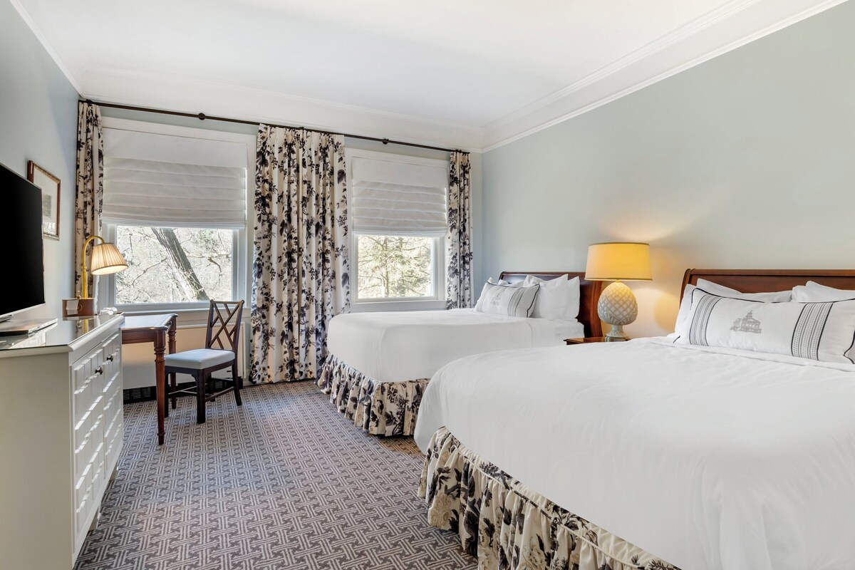 A hotel room with two beds, a desk, a chair, and a television.
