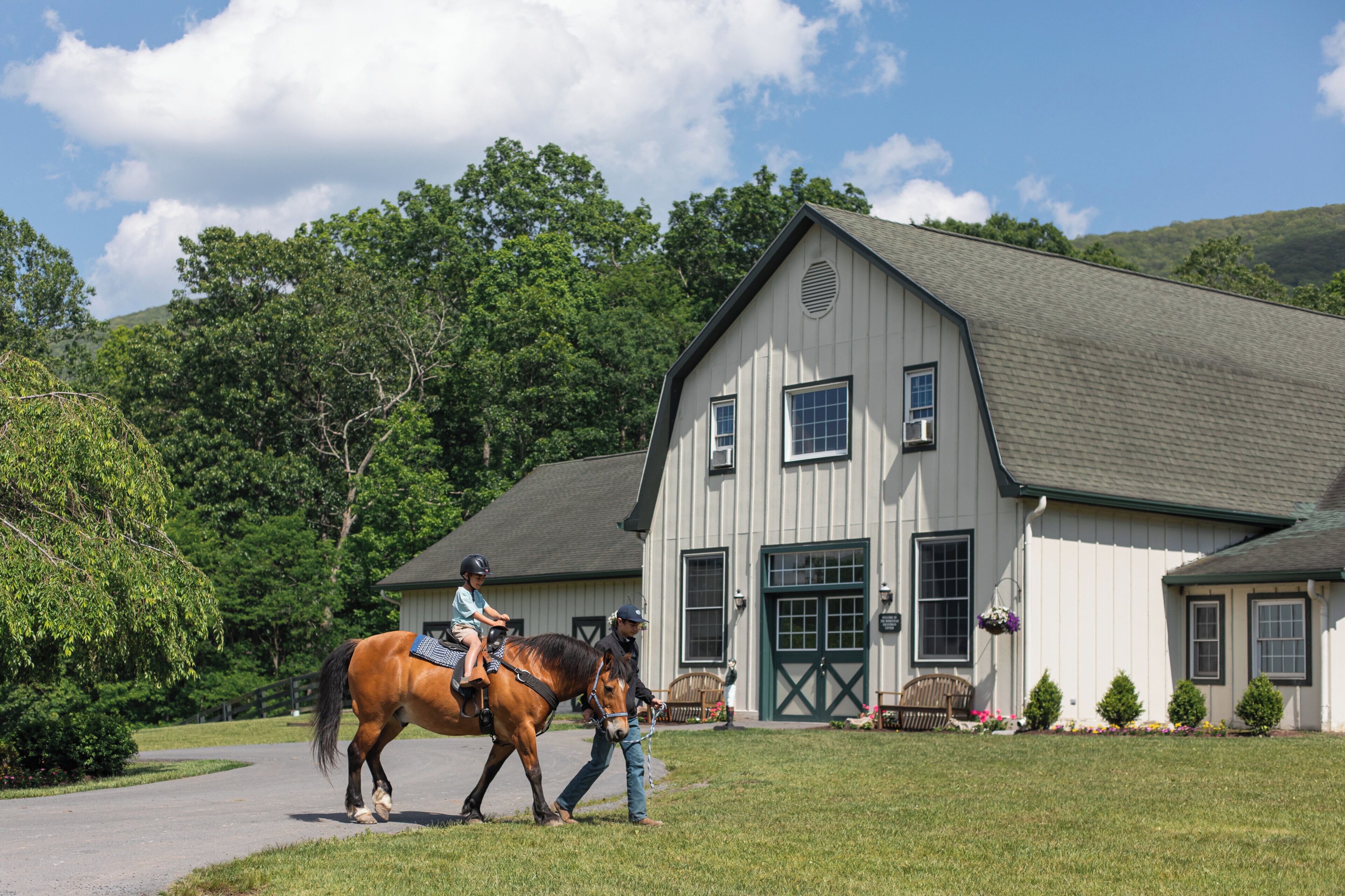 A stable with a horse and rider, a large white building with a green door, and a well-maintained lawn.