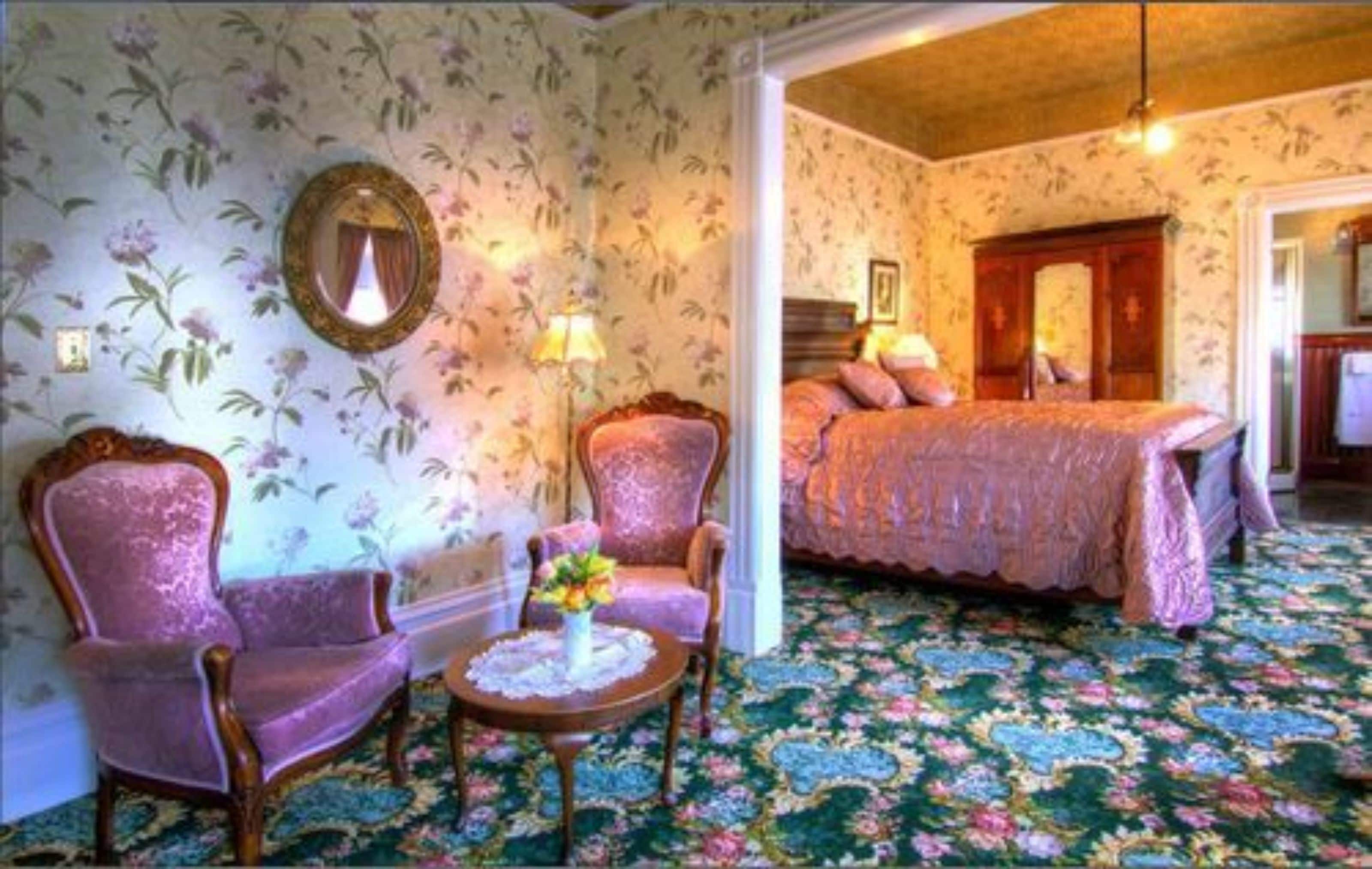 A room with floral wallpaper, a bed with a pink bedspread, two armchairs, a small table with a vase, and a mirror on the wall.