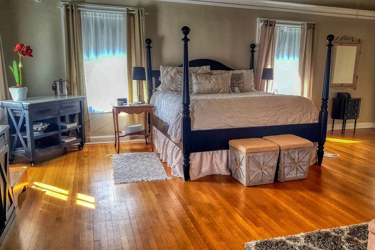 A spacious bedroom with a large bed, a nightstand, a dresser, and a window with curtains.