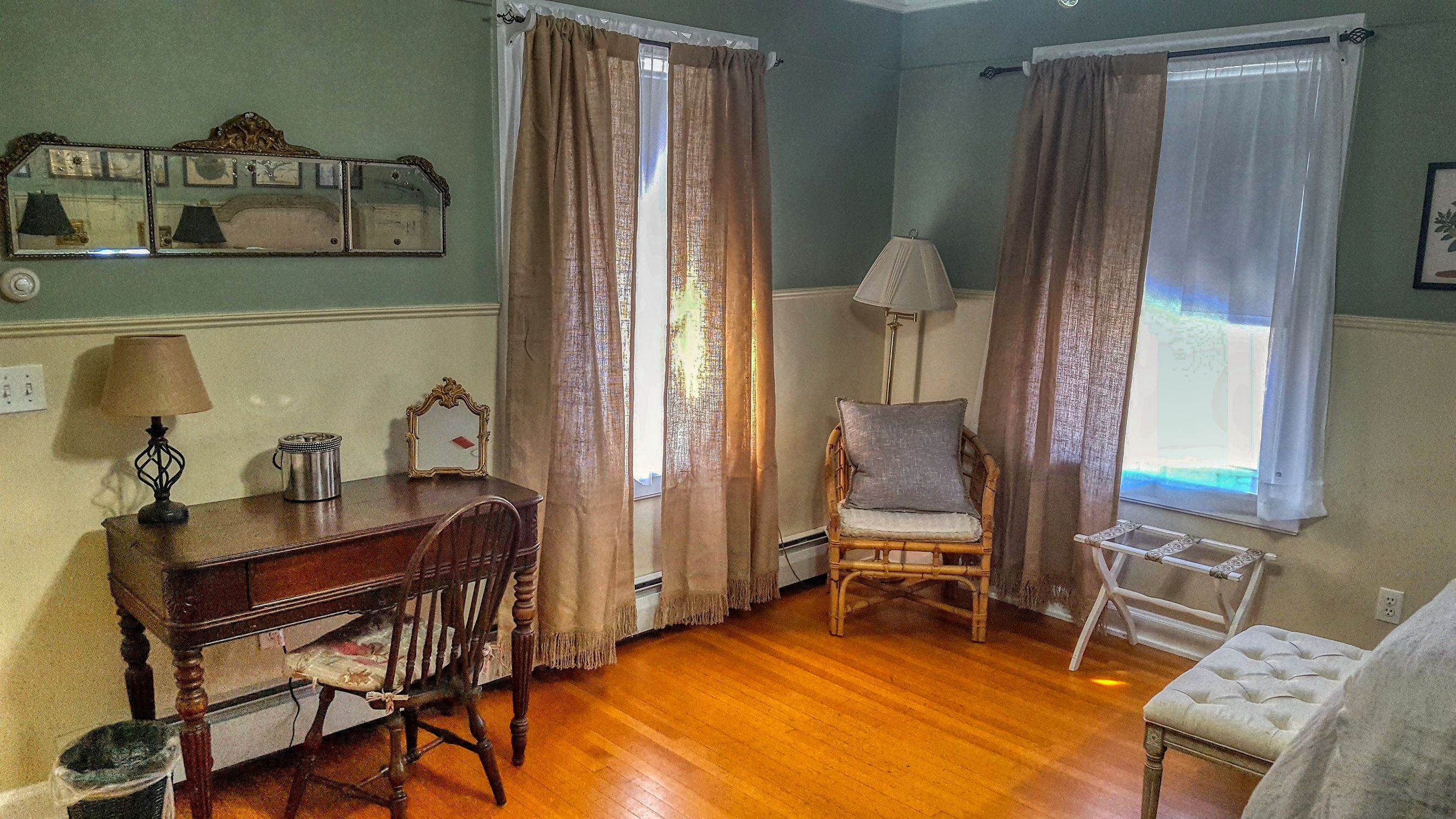 A room with wooden flooring, a desk with a lamp, a chair, and two windows with curtains.