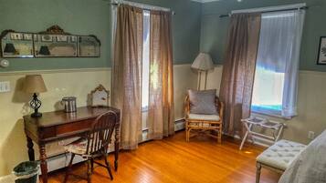 A room with wooden flooring, a desk with a lamp, a chair, and two windows with curtains.