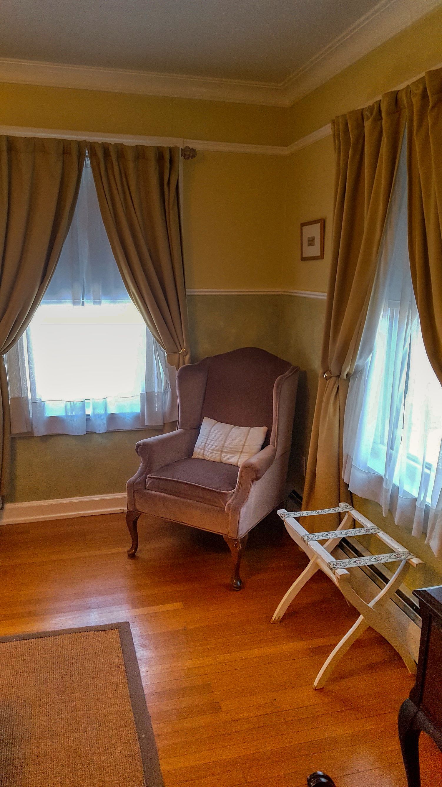 A room with a wooden floor, a brown armchair with a cushion, a white folding chair, and a window with curtains.