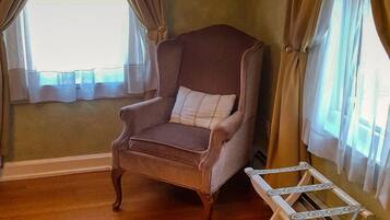 A room with a wooden floor, a brown armchair with a cushion, a white folding chair, and a window with curtains.