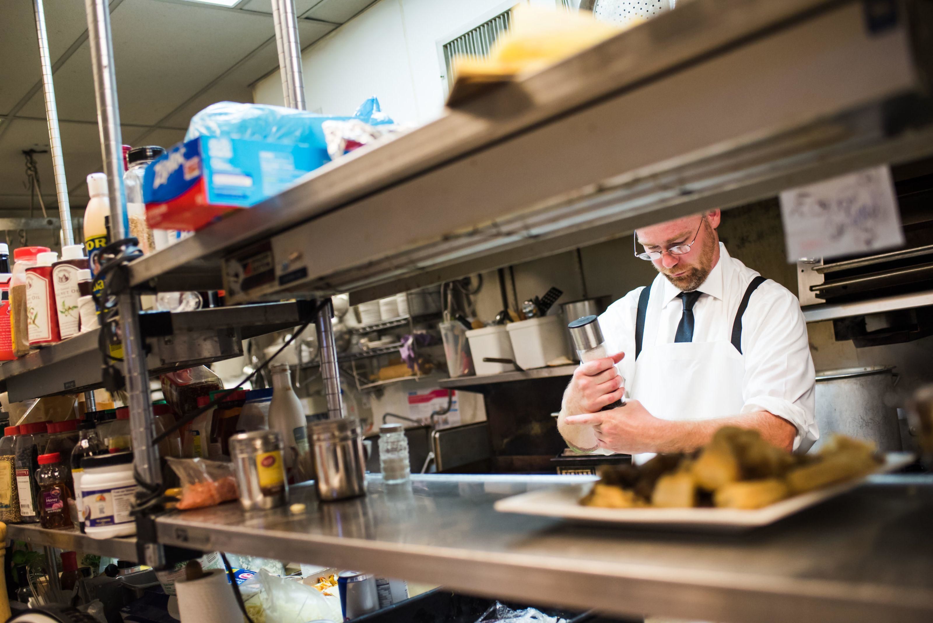 Un chef dans une cuisine professionnelle, avec des surfaces en acier inoxydable, divers ustensiles de cuisine et des ingrédients.