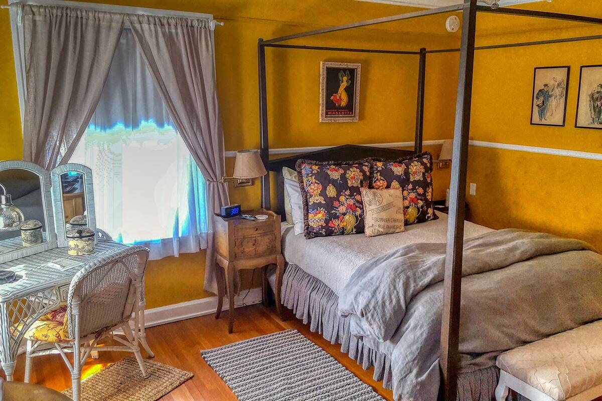 A four-poster bed with floral pillows, a bedside table with a lamp, a mirror, and a small clock.