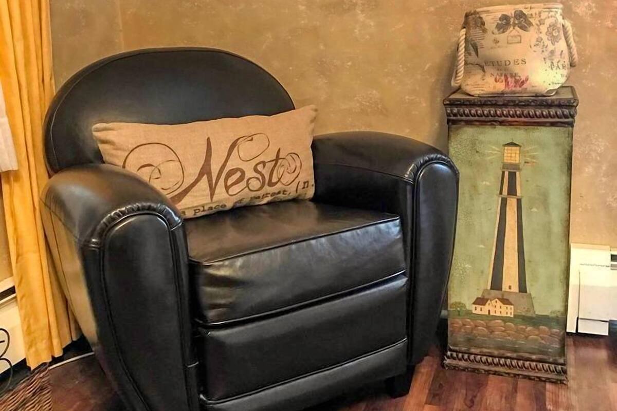 A black leather armchair with a cushion labeled "Nest" in a room with wooden flooring, a wall with framed pictures, and a decorative lamp with a lighthouse design.