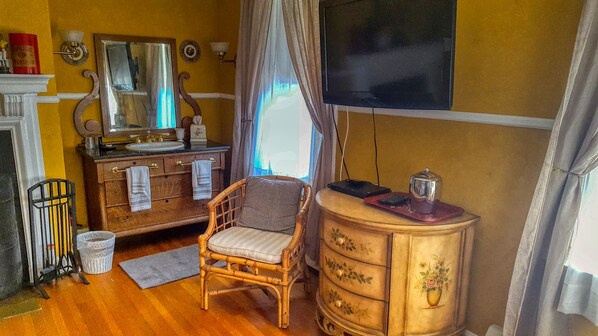 A room with a wooden dresser, a wicker chair, a small table with a lamp, and a mirror above the dresser. - The Lilac Inn (Brandon)