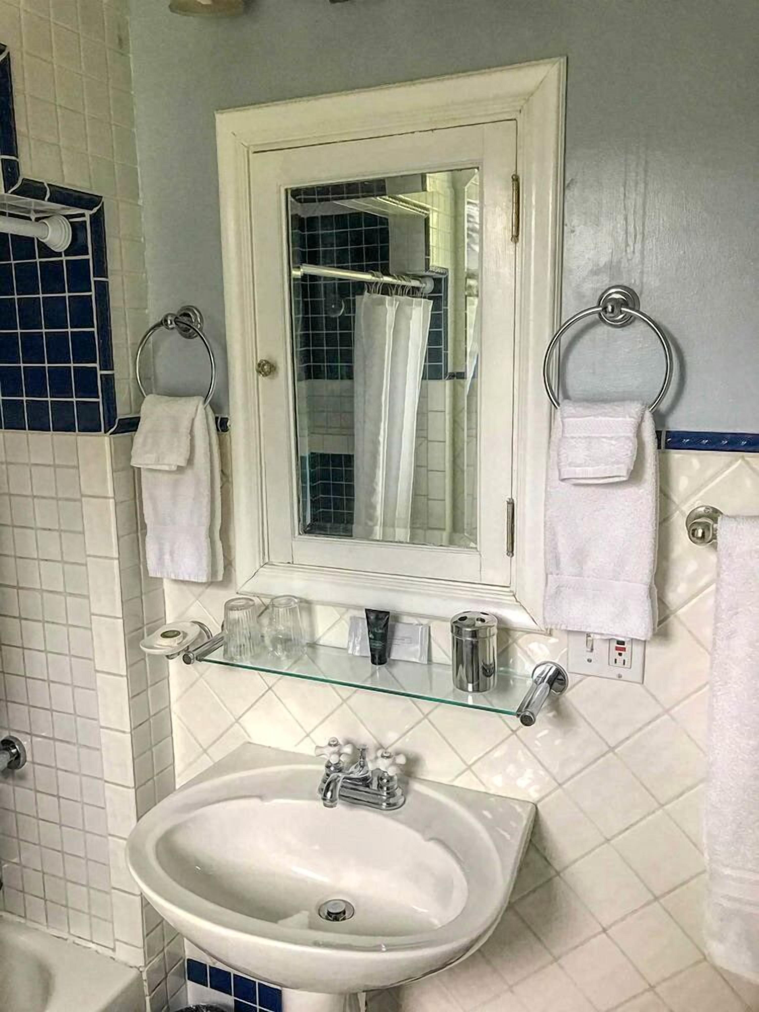 Une salle de bain avec un lavabo blanc, une étagère en verre où sont rangés les articles de toilette, et un miroir au-dessus du lavabo.