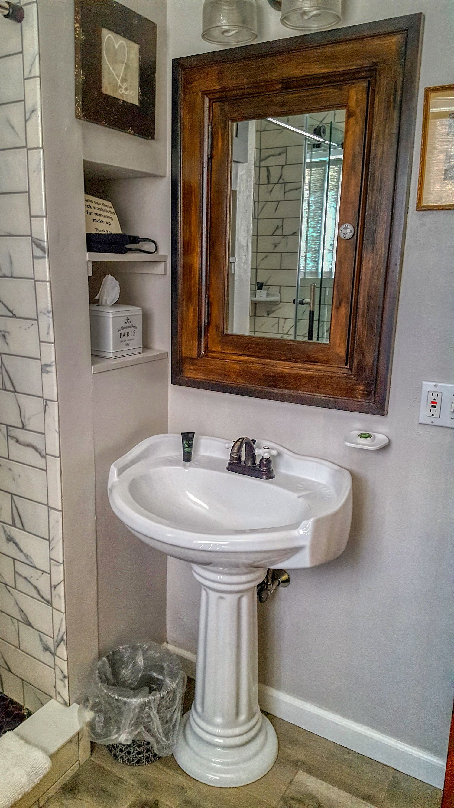 Un baño con lavamanos blanco de pedestal, espejo con marco de madera y pared de ducha con diseño de mármol.